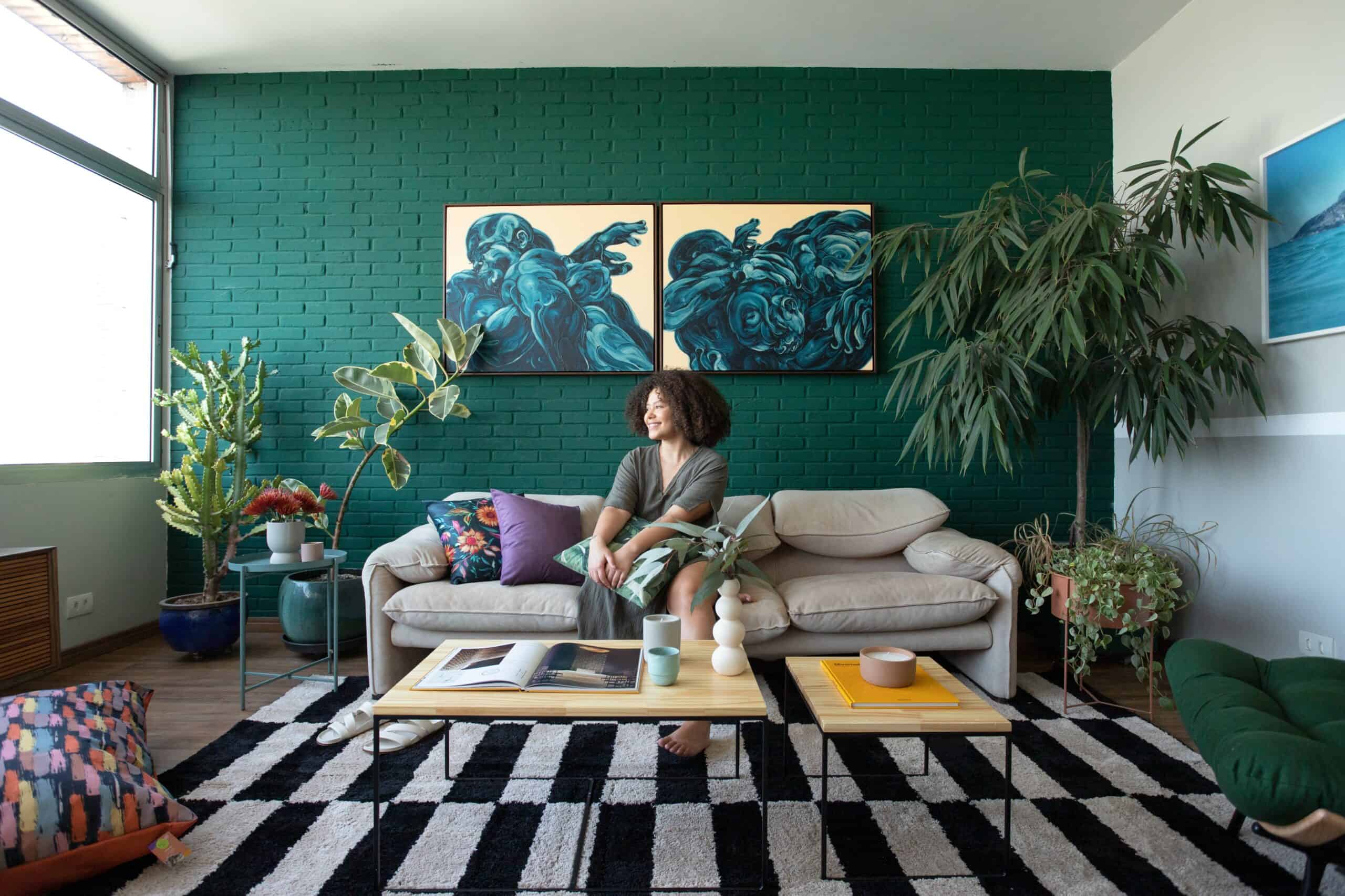 modelo sentada em uma sala repleta de plantas, no espaço tem tapete, sofá, mesa de centro e quadros