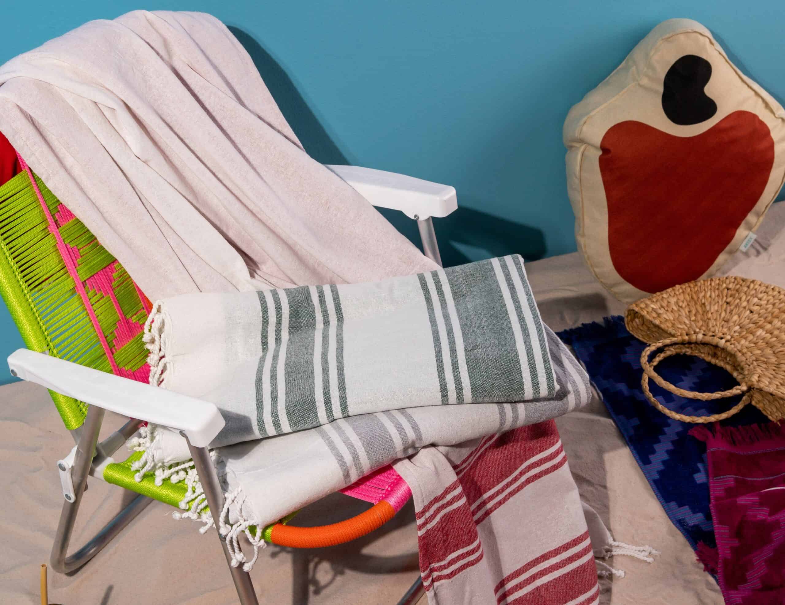 quatro toalhas de praia sobre uma cadeira, ao lado almofada em forma de caju e uma bolsa de palha