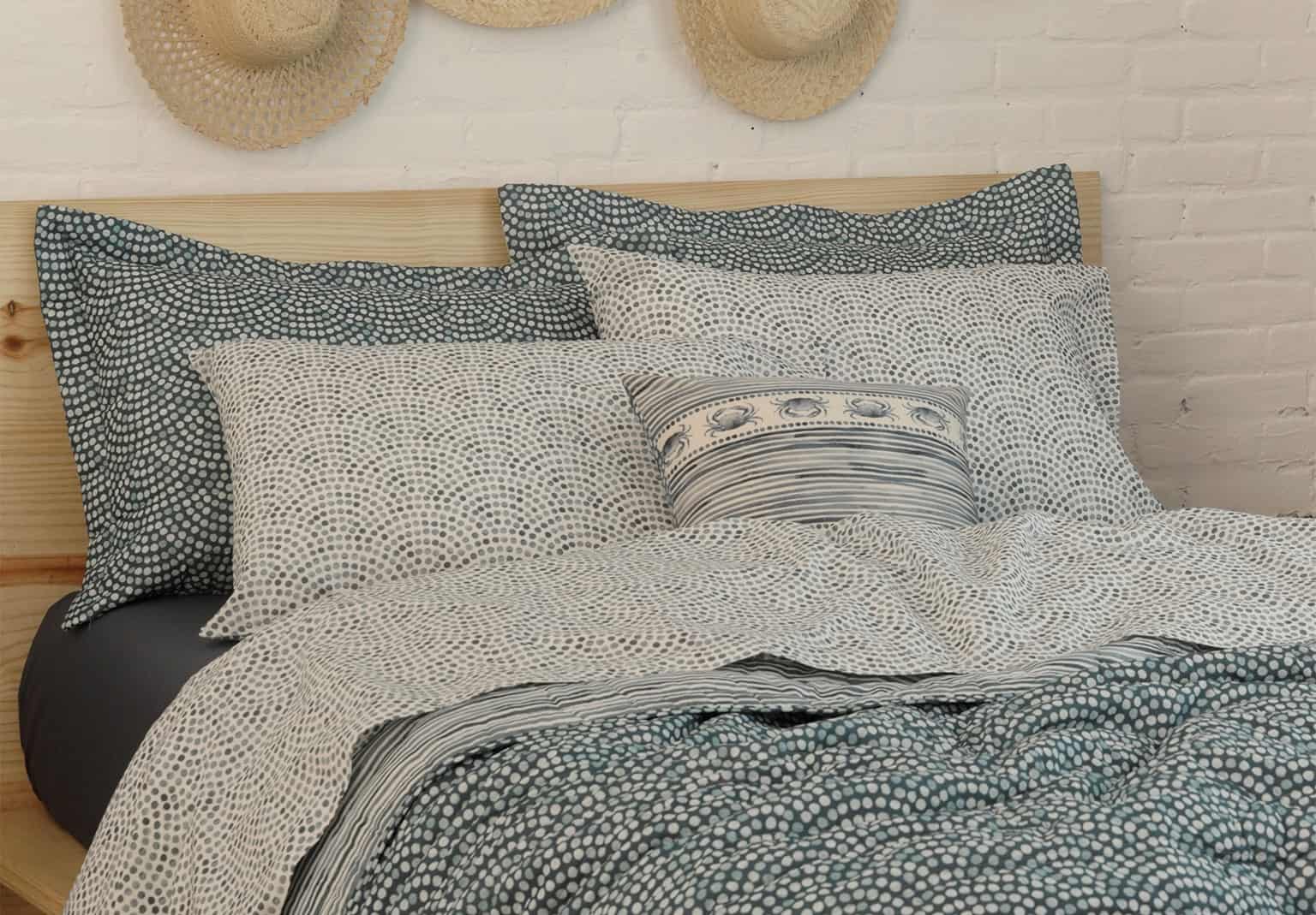 uma cama com uma estampa azul que refaz as ondas do mar de um jeito encantador, há quatro travesseiros e uma almofada, no fundo, chapeis de palha decorativos