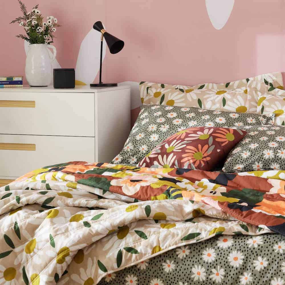 um quarto temático com margaridas: na cama a roupa de cama possui essa estampa, a parede de fundo tem desenhos com o tema e na mesa de cabeceira tem um jarro com as flores 